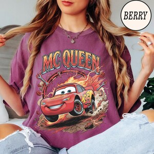 Puede incluir: Una camiseta color baya con un estampado gr&aacute;fico de un coche de carreras rojo con el n&uacute;mero 95, rodeado de llamas, herramientas y escombros. El texto MC QUEEN est&aacute; impreso en una fuente negrita y arqueada sobre la ilustraci&oacute;n del coche.