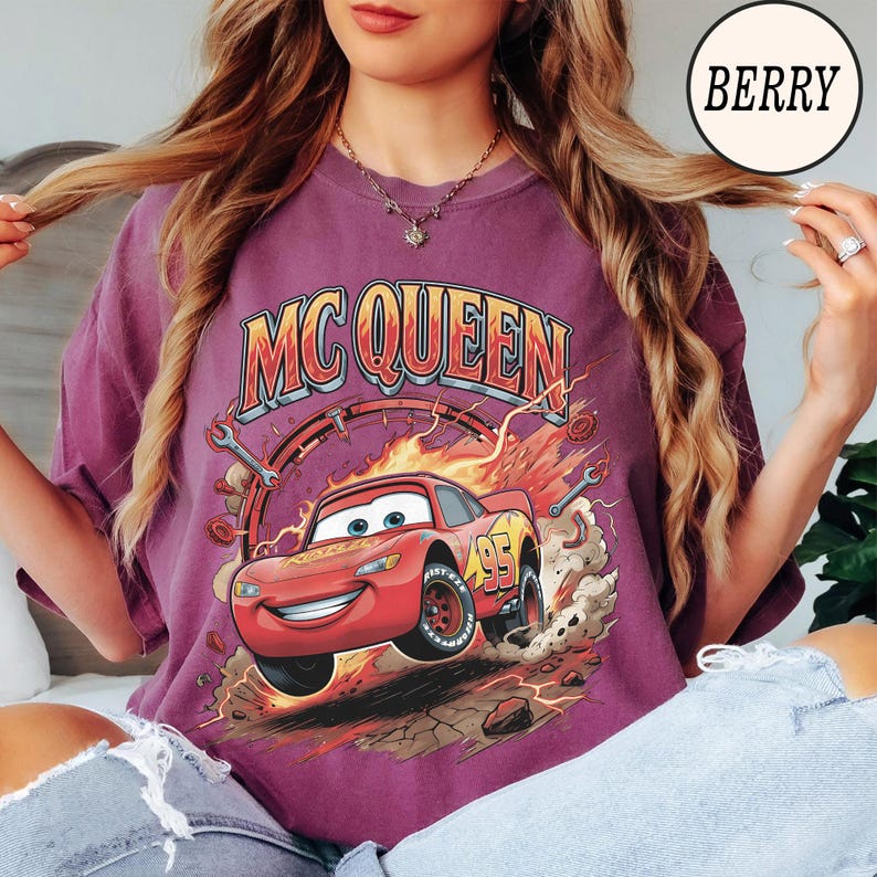 Puede incluir: Una camiseta color baya con un estampado gr&aacute;fico de un coche de carreras rojo con el n&uacute;mero 95, rodeado de llamas, herramientas y escombros. El texto MC QUEEN est&aacute; impreso en una fuente negrita y arqueada sobre la ilustraci&oacute;n del coche.