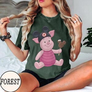 May include: Dark green t-shirt featuring a cartoon Piglet character wearing a Mickey Mouse hat and holding a chocolate-covered ice cream bar. The shirt has a round neckline and short sleeves. The word "FOREST" is printed on a white circle.