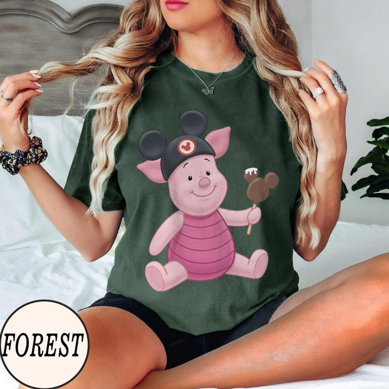 May include: Dark green t-shirt featuring a cartoon Piglet character wearing a Mickey Mouse hat and holding a chocolate-covered ice cream bar. The shirt has a round neckline and short sleeves. The word "FOREST" is printed on a white circle.