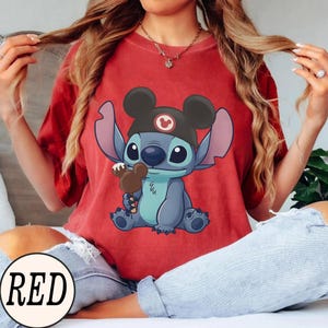 K&ouml;nnte beinhalten: Rotes T-Shirt mit einer Cartoon-Figur, Stitch, die einen Mickey Mouse-Hut tr&auml;gt und ein Schokoladeneis h&auml;lt. Das Wort "RED" ist in einem wei&szlig;en Kreis unten aufgedruckt. Das T-Shirt hat eine einfarbige rote Farbe.