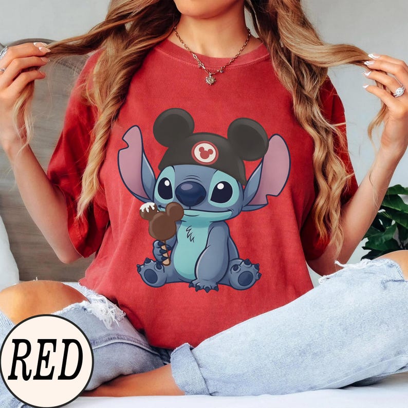 K&ouml;nnte beinhalten: Rotes T-Shirt mit einer Cartoon-Figur, Stitch, die einen Mickey Mouse-Hut tr&auml;gt und ein Schokoladeneis h&auml;lt. Das Wort "RED" ist in einem wei&szlig;en Kreis unten aufgedruckt. Das T-Shirt hat eine einfarbige rote Farbe.