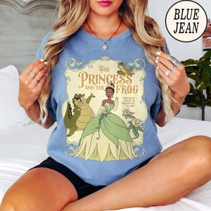 Op de afbeelding: Een blauw t-shirt met ronde hals en een grafische print van Tiana uit The Princess and the Frog, samen met Louis de alligator en een kleine kikker. De tekst luidt The Princess and the Frog en The Real Magic is Believing in You in een kader.