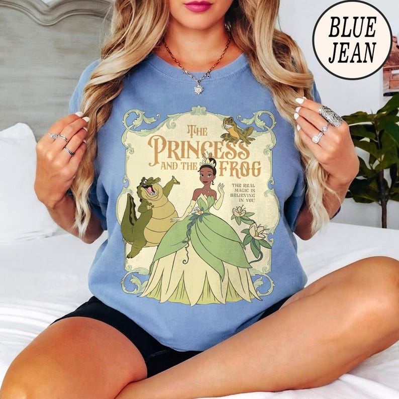 Op de afbeelding: Een blauw t-shirt met ronde hals en een grafische print van Tiana uit The Princess and the Frog, samen met Louis de alligator en een kleine kikker. De tekst luidt The Princess and the Frog en The Real Magic is Believing in You in een kader.