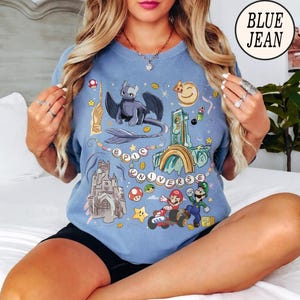 May include: Light blue t-shirt with a colorful graphic design featuring a dragon, castle, and video game characters. The words "EPIC UNIVERSE" are also displayed. A circular logo in the upper right corner reads "BLUE JEAN".