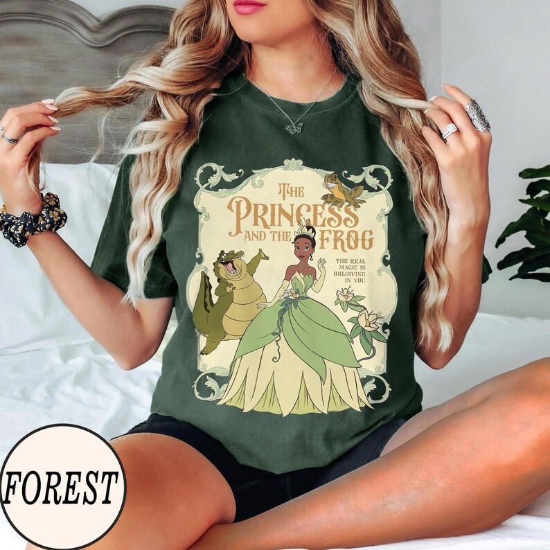 Op de afbeelding: Een donkergroen t-shirt met een grafische print van Tiana uit The Princess and the Frog, samen met Louis de alligator en Ray de vuurvlieg. De tekst luidt The Princess and the Frog en The Real Magic Is Believing In You.