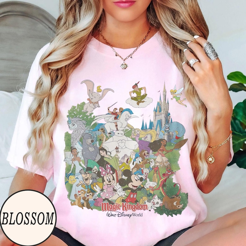 Peut inclure: T-shirt rose clair avec un motif color&eacute; de personnages Disney, dont Mickey Mouse, Dumbo et Peter Pan, avec le texte "Magic Kingdom Walt Disney World". Le mot "BLOSSOM" est dans un cercle blanc en bas &agrave; gauche.