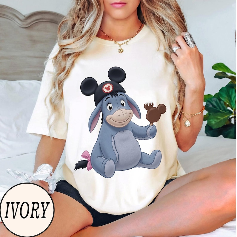 May include: Ivory-colored t-shirt featuring a cartoon illustration of Eeyore wearing a Mickey Mouse hat and holding a Mickey-shaped ice cream bar. The text "IVORY" is in a circle at the bottom. The design is centered on the shirt.