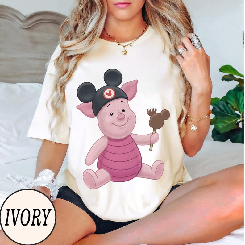 May include: Ivory-colored t-shirt featuring a cartoon Piglet character wearing a Mickey Mouse hat and holding a chocolate-covered ice cream bar. The character is pink with a striped pink shirt. The word "IVORY" is printed on the bottom left.