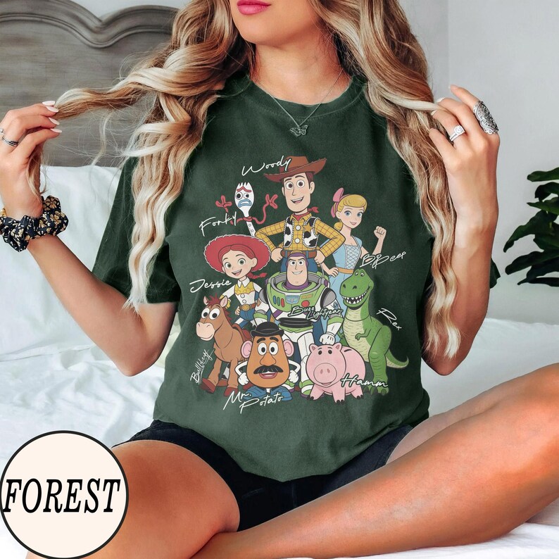 May include: Dark green t-shirt featuring a graphic of Toy Story characters, including Woody, Buzz Lightyear, and Jessie. The word "FOREST" is printed on a white circle at the bottom. The shirt is a comfortable, casual style.