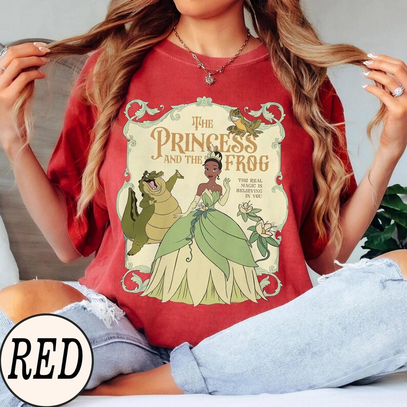 Op de afbeelding: Een rood t-shirt met een grafische print van Tiana uit The Princess and the Frog, een groene alligator en een kikker. Het ontwerp bevat sierlijke randen, florale accenten en de tekst The Princess and the Frog en The Real Magic Is Believing In You.