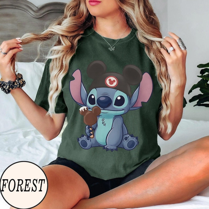 K&ouml;nnte beinhalten: Dunkelgr&uuml;nes T-Shirt mit einer Cartoonfigur, die einen Mickey Mouse-Hut tr&auml;gt und ein Schokoladeneis h&auml;lt. Die Figur ist blau mit gro&szlig;en Augen und rosa Ohren. Das Wort "FOREST" ist auf einem wei&szlig;en Kreis aufgedruckt.