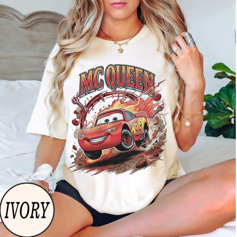 Puede incluir: Una camiseta de manga corta color marfil con un estampado gr&aacute;fico del personaje de coche de carreras rojo Lightning McQueen y el texto MC QUEEN encima. El dise&ntilde;o incluye llamas, llaves inglesas y el n&uacute;mero 95 en el coche sobre un fondo polvoriento.