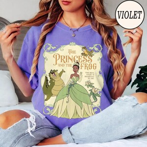 Op de afbeelding: Een paars t-shirt met een grafische print van The Princess and the Frog. Het ontwerp bevat een centrale illustratie van een prinses in een groene jurk, een groene kikker en een lachende alligator, omringd door sierlijk krulwerk en tekst.
