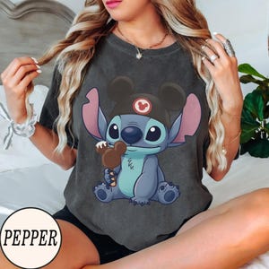 K&ouml;nnte beinhalten: Dunkelgraues T-Shirt mit einer Cartoon-Figur Stitch, die einen Mickey Mouse-Hut tr&auml;gt und ein Schokoladeneis h&auml;lt. Die Figur ist blau mit gro&szlig;en Ohren und Augen. Das T-Shirt hat einen Used-Look.