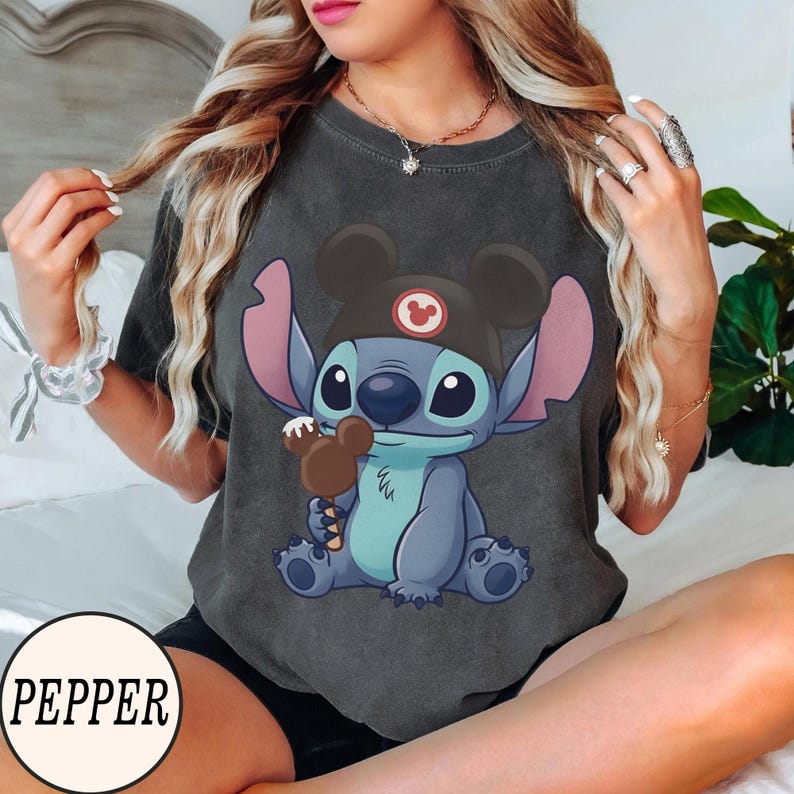 K&ouml;nnte beinhalten: Dunkelgraues T-Shirt mit einer Cartoon-Figur Stitch, die einen Mickey Mouse-Hut tr&auml;gt und ein Schokoladeneis h&auml;lt. Die Figur ist blau mit gro&szlig;en Ohren und Augen. Das T-Shirt hat einen Used-Look.