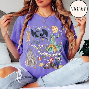 May include: Violet-colored t-shirt featuring a graphic design with a dragon, castle, and video game characters. The text "EPIC UNIVERSE" is displayed. The shirt has short sleeves and a crew neck.