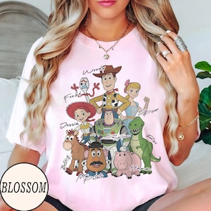 May include: Pink t-shirt featuring a colorful graphic of characters from the animated movie Toy Story. The design includes Woody, Buzz Lightyear, Jessie, and other beloved characters. The word "Blossom" is printed at the bottom left.