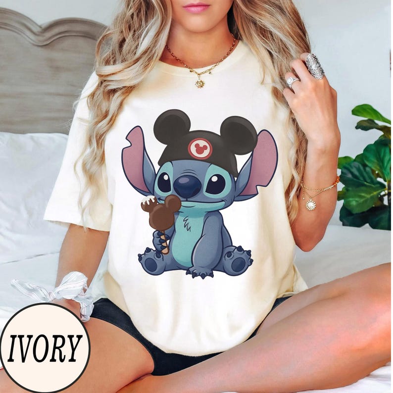 K&ouml;nnte beinhalten: Elfenbeinfarbenes T-Shirt mit einer Cartoon-Figur Stitch, die einen Mickey Mouse-Hut tr&auml;gt und einen Schokoladeneisriegel h&auml;lt. Das Design enth&auml;lt das Wort "IVORY" in einem kreisf&ouml;rmigen Schriftzug unten links.