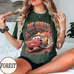 Puede incluir: Una camiseta verde oscuro con un gr&aacute;fico de un coche de carreras rojo con el n&uacute;mero 95, rodeado de llamas, humo y herramientas. El texto MC QUEEN est&aacute; impreso en una fuente negrita y arqueada sobre el coche, que tiene ojos grandes.
