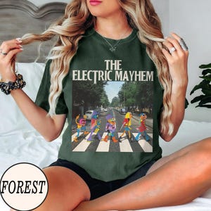 Pode incluir: T-shirt verde escuro com gr&aacute;fico "The Electric Mayhem". O gr&aacute;fico mostra personagens dos Muppets atravessando uma rua. A camiseta tem um corte folgado.