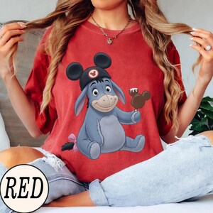 May include: A red t-shirt featuring a cartoon illustration of Eeyore wearing a Mickey Mouse hat and holding a Mickey-shaped ice cream bar. The word "RED" is in a white circle at the bottom.