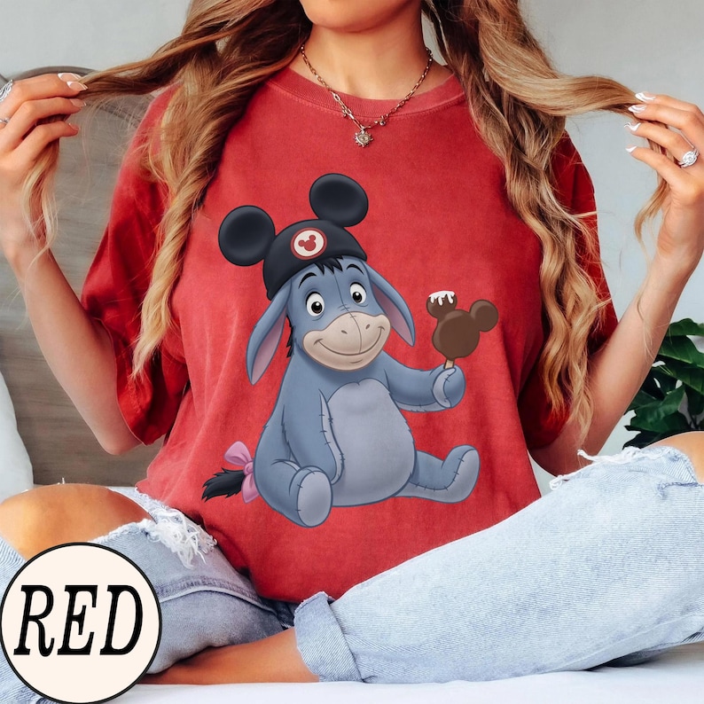 May include: A red t-shirt featuring a cartoon illustration of Eeyore wearing a Mickey Mouse hat and holding a Mickey-shaped ice cream bar. The word "RED" is in a white circle at the bottom.
