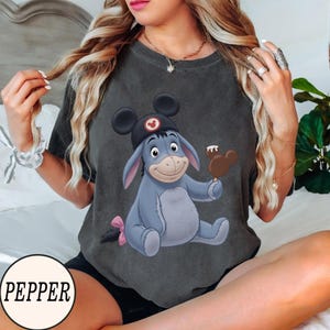 May include: Dark gray t-shirt featuring a cartoon illustration of Eeyore wearing a Mickey Mouse hat and holding a Mickey Mouse-shaped ice cream bar. The shirt has a distressed look and a pink bow on Eeyore's tail.