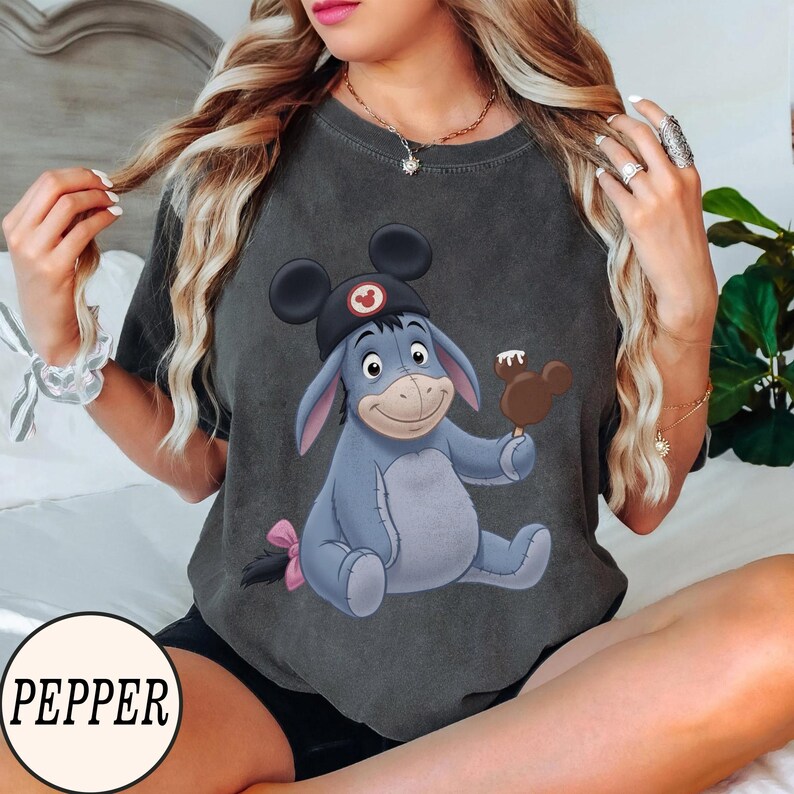 May include: Dark gray t-shirt featuring a cartoon illustration of Eeyore wearing a Mickey Mouse hat and holding a Mickey Mouse-shaped ice cream bar. The shirt has a distressed look and a pink bow on Eeyore's tail.