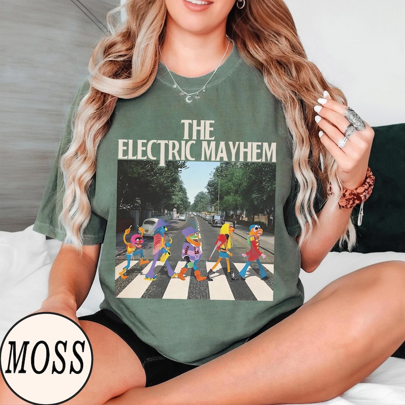 Pode incluir: T-shirt verde com gr&aacute;fico "The Electric Mayhem". O design &eacute; uma par&oacute;dia da capa do &aacute;lbum Abbey Road dos Beatles, com personagens dos Muppets atravessando uma faixa de pedestres. A camiseta tem mangas curtas e gola redonda.