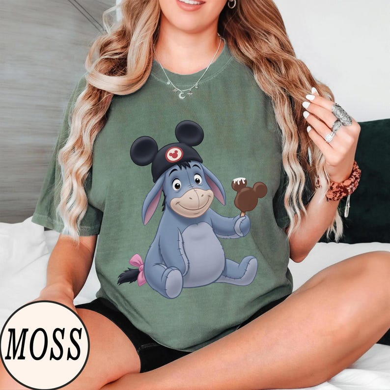 May include: Sage green t-shirt featuring a cartoon Eeyore character wearing a Mickey Mouse hat and holding a Mickey Mouse-shaped ice cream bar. The character is light blue with a pink bow on its tail. The shirt has the word "MOSS" in a circle.