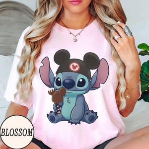 K&ouml;nnte beinhalten: Hellrosa T-Shirt mit einer Cartoon-Illustration von Stitch, der einen Mickey Mouse-Hut tr&auml;gt und einen Schokoladeneis am Stiel h&auml;lt. Das Wort "BLOSSOM" ist in einem wei&szlig;en Kreis unten links aufgedruckt.