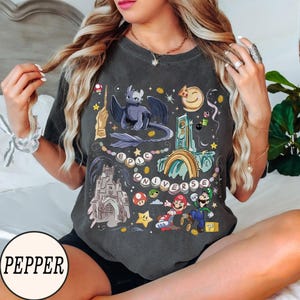 May include: Dark gray t-shirt with a colorful graphic featuring a dragon, castle, and video game characters. The words "EPIC UNIVERSE" are printed on the shirt. The shirt has a crew neck and short sleeves.