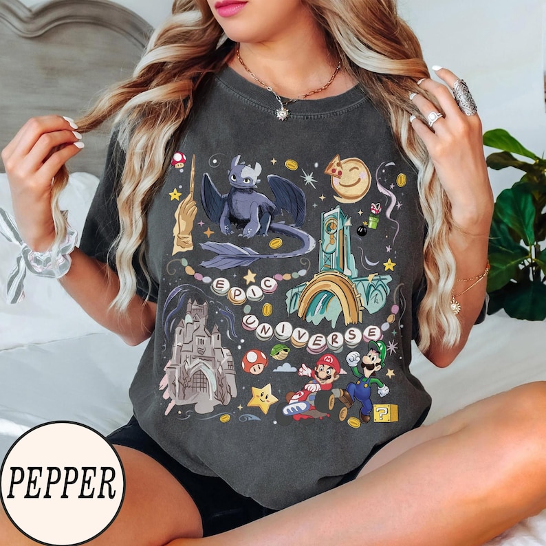May include: Dark gray t-shirt with a colorful graphic featuring a dragon, castle, and video game characters. The words "EPIC UNIVERSE" are printed on the shirt. The shirt has a crew neck and short sleeves.