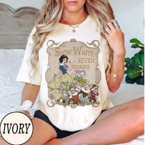 Op de afbeelding: Een ivoorkleurig t-shirt met korte mouwen en een vintage-stijl afbeelding van Sneeuwwitje en de zeven dwergen in een sierlijk gouden kader. De tekst luidt Snow White and Seven Dwarfs. Het ontwerp toont de personages in klassieke kleuren.