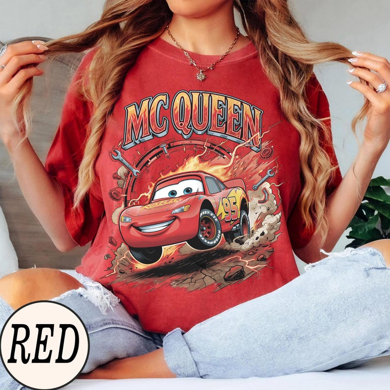 Puede incluir: Una camiseta roja de manga corta con un estampado gr&aacute;fico de un coche de carreras rojo con el n&uacute;mero 95 en el lateral. El texto MC QUEEN aparece en letras grandes sobre el coche, rodeado de llamas, nubes de polvo y herramientas.