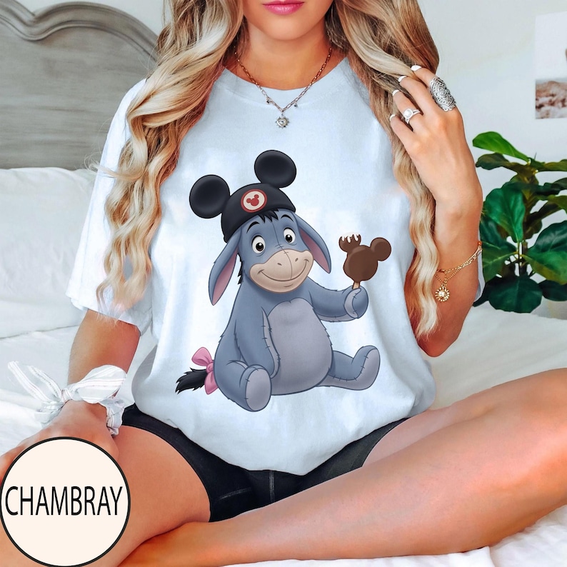 May include: White t-shirt featuring a cartoon illustration of Eeyore wearing a Mickey Mouse hat and holding a Mickey Mouse-shaped ice cream bar. Eeyore is light blue with a pink bow on his tail. The shirt has a round neckline.