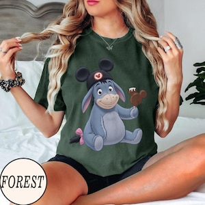 May include: Forest green t-shirt featuring a cartoon illustration of Eeyore wearing a Mickey Mouse hat and holding a Mickey Mouse-shaped ice cream bar. The shirt has a round neck and short sleeves. The word "FOREST" is printed on the bottom left.