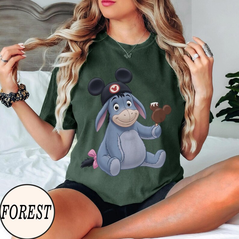 May include: Forest green t-shirt featuring a cartoon illustration of Eeyore wearing a Mickey Mouse hat and holding a Mickey Mouse-shaped ice cream bar. The shirt has a round neck and short sleeves. The word "FOREST" is printed on the bottom left.