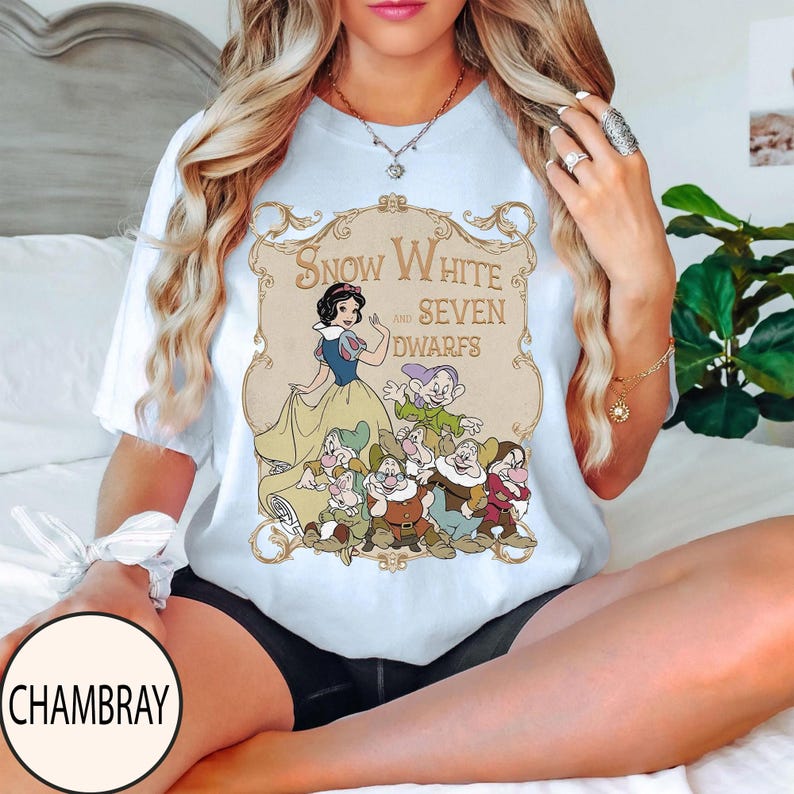 Op de afbeelding: Een lichtblauw t-shirt met een grafische print van Sneeuwwitje en de zeven dwergen in een sierlijk goudkleurig kader. De tekst "Snow White and Seven Dwarfs" staat in een decoratief lettertype boven de personages op het t-shirt.