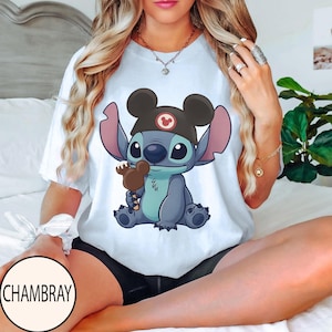 K&ouml;nnte beinhalten: Wei&szlig;es T-Shirt mit einer Cartoon-Illustration von Stitch, der einen Mickey Mouse-Hut tr&auml;gt und ein braunes Eis h&auml;lt. Das Design ist zentriert. Der Markenname "CHAMBRAY" ist auf einem wei&szlig;en Kreis unten links aufgedruckt.