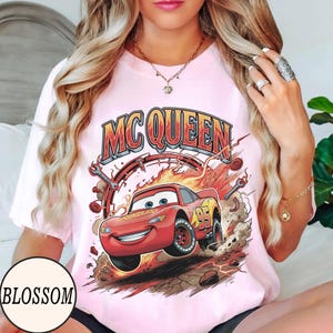 Puede incluir: Una camiseta rosa claro con un gr&aacute;fico de un coche de carreras rojo con el n&uacute;mero 95 en el lateral, rodeado de llamas, polvo y herramientas. El texto MC QUEEN est&aacute; impreso en letras grandes, negritas, naranjas y rojas sobre la ilustraci&oacute;n.
