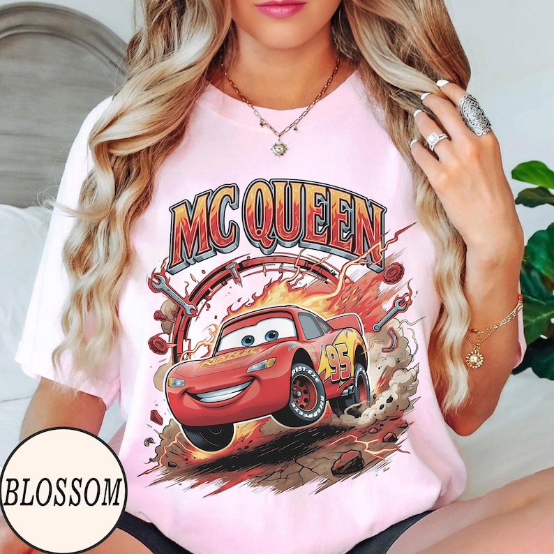 Puede incluir: Una camiseta rosa claro con un gr&aacute;fico de un coche de carreras rojo con el n&uacute;mero 95 en el lateral, rodeado de llamas, polvo y herramientas. El texto MC QUEEN est&aacute; impreso en letras grandes, negritas, naranjas y rojas sobre la ilustraci&oacute;n.