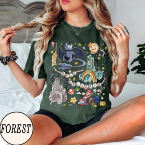 May include: Dark green t-shirt with a colorful graphic featuring a dragon, castle, rainbow, and video game characters. The text "EPIC UNIVERSE" is also displayed. The shirt has a round neckline and short sleeves.