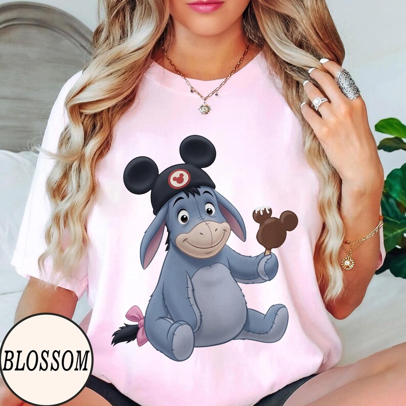 May include: Pink t-shirt featuring a cartoon illustration of Eeyore wearing a Mickey Mouse hat and holding a Mickey Mouse ice cream bar. The word "BLOSSOM" is printed in a white circle at the bottom left.