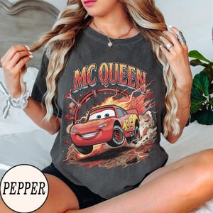 Puede incluir: Una camiseta de manga corta gris carb&oacute;n con un estampado gr&aacute;fico de un coche de carreras rojo con el n&uacute;mero 95 en el lateral. El texto MC QUEEN est&aacute; impreso en letras grandes amarillas y rojas sobre el coche, rodeado de llamas, llaves inglesas y polvo.