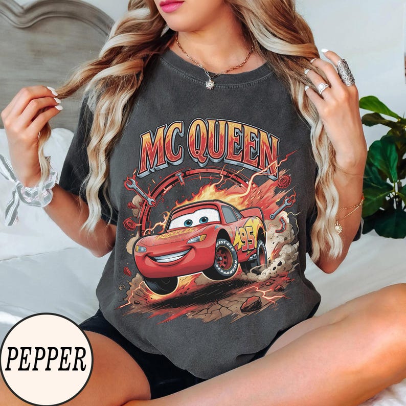 Puede incluir: Una camiseta de manga corta gris carb&oacute;n con un estampado gr&aacute;fico de un coche de carreras rojo con el n&uacute;mero 95 en el lateral. El texto MC QUEEN est&aacute; impreso en letras grandes amarillas y rojas sobre el coche, rodeado de llamas, llaves inglesas y polvo.