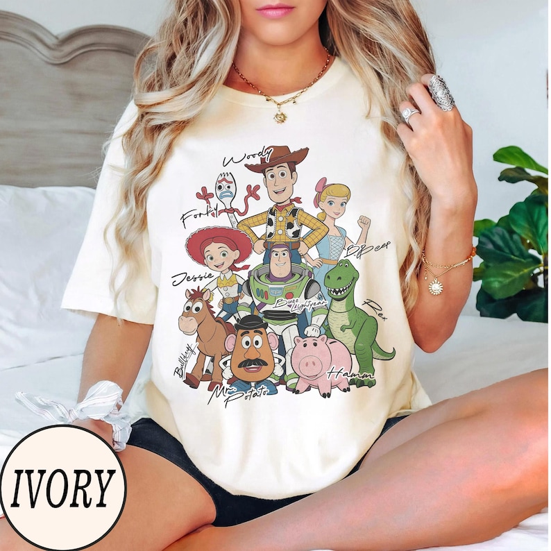 May include: Ivory-colored t-shirt featuring a graphic of characters from the animated film Toy Story. The design includes Woody, Buzz Lightyear, Jessie, and other characters with their names written above them. The word "IVORY" is printed in a circle at the bottom.