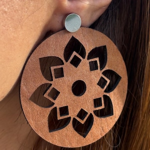 May include: Copper-colored leather earrings with a laser-cut floral design. The earrings are circular with a silver stud at the top. The floral pattern is black and features a central dot surrounded by stylized petals and geometric shapes.