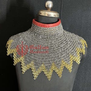 Könnte beinhalten: Ein Kettenkragen, der auf einer Schaufensterpuppe ausgestellt ist. Der Kragen hat einen silberfarbenen Kettenkörper mit einem goldfarbenen, gewellten Rand. Ein roter Lederbesatz säumt die Halsöffnung. Die Schaufensterpuppe hat ein Metall-Halsstück.